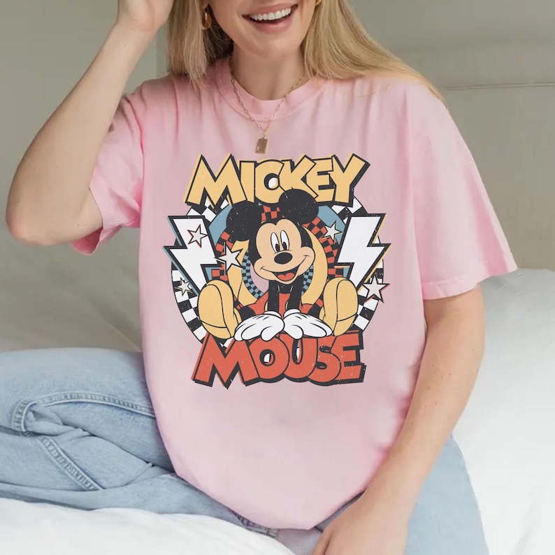 May include: Pink t-shirt featuring a vintage Mickey Mouse graphic. The design includes Mickey Mouse sitting with the words "Mickey Mouse" in a retro font, surrounded by stars and lightning bolts. The shirt is a light pink color.