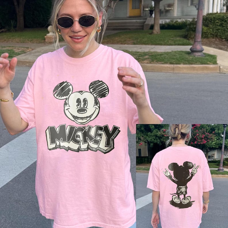 May include: A light pink t-shirt featuring a black and white graphic of Mickey Mouse with the word "Mickey" in a bold font. The back of the shirt shows a silhouette of Mickey Mouse with his arms raised in a peace sign.