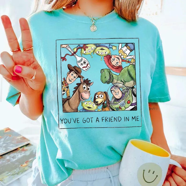 K&ouml;nnte beinhalten: Ein hellblaues T-Shirt mit einer gerahmten Grafik von Toy Story-Charakteren, darunter Woody, Buzz Lightyear und Rex. Der Text "YOU'VE GOT A FRIEND IN ME" ist darunter gedruckt. Eine wei&szlig;e Tasse mit gelbem Innenraum und Smiley ist ebenfalls zu sehen.