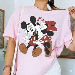 May include: Light pink t-shirt featuring a graphic of Mickey Mouse and Minnie Mouse embracing, with hearts above them. Mickey is wearing red shorts and brown shoes, while Minnie wears a red polka-dot skirt and bow. The shirt is a casual style.
