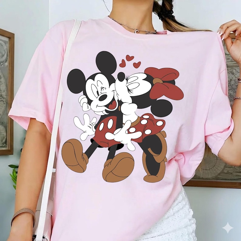 May include: Light pink t-shirt featuring a graphic of Mickey Mouse and Minnie Mouse embracing, with hearts above them. Mickey is wearing red shorts and brown shoes, while Minnie wears a red polka-dot skirt and bow. The shirt is a casual style.