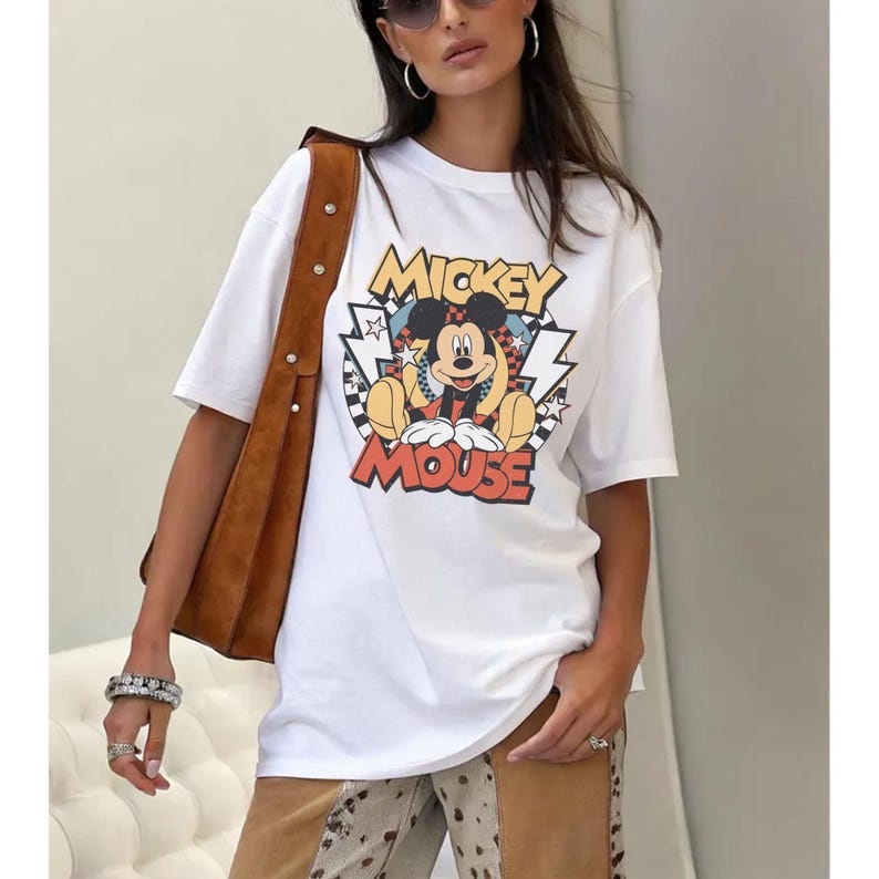 May include: White t-shirt featuring a colorful graphic of Mickey Mouse with the text "Mickey Mouse". The design includes a checkered background and lightning bolt accents. The shirt has short sleeves and a crew neck.