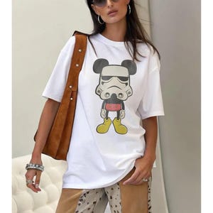 May include: White t-shirt with a cartoon design combining Mickey Mouse and a Stormtrooper. The graphic uses black, white, red, and yellow. The tee has short sleeves and a round neck.