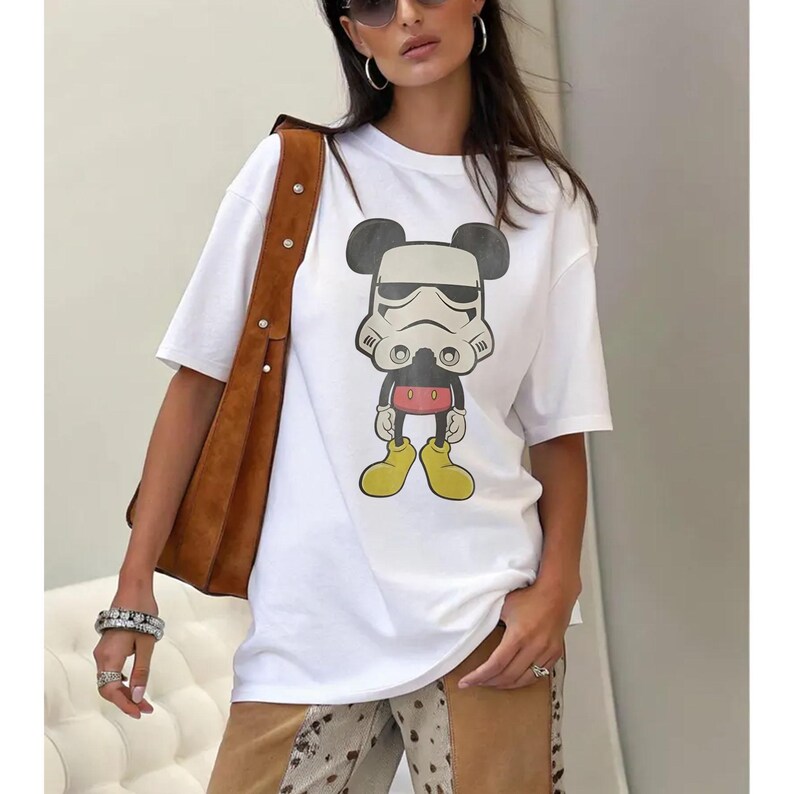 May include: White t-shirt with a cartoon design combining Mickey Mouse and a Stormtrooper. The graphic uses black, white, red, and yellow. The tee has short sleeves and a round neck.