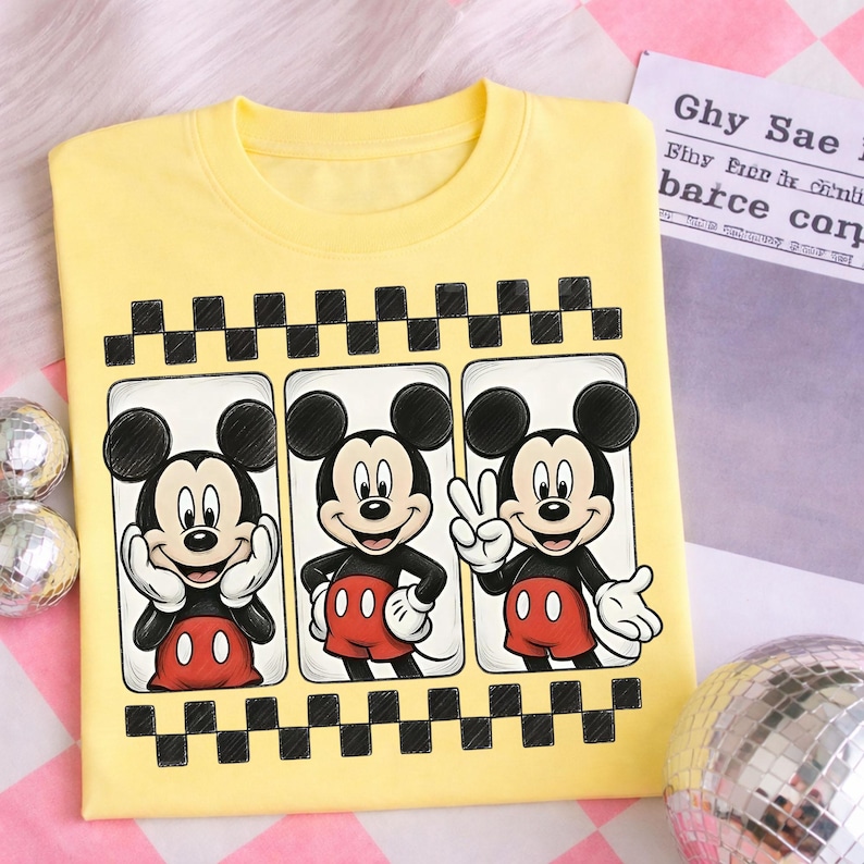 May include: Yellow t-shirt featuring three panels with Mickey Mouse illustrations. Each panel shows Mickey in a different pose, with a black and white checkered border. The shirt has a crew neck and short sleeves.