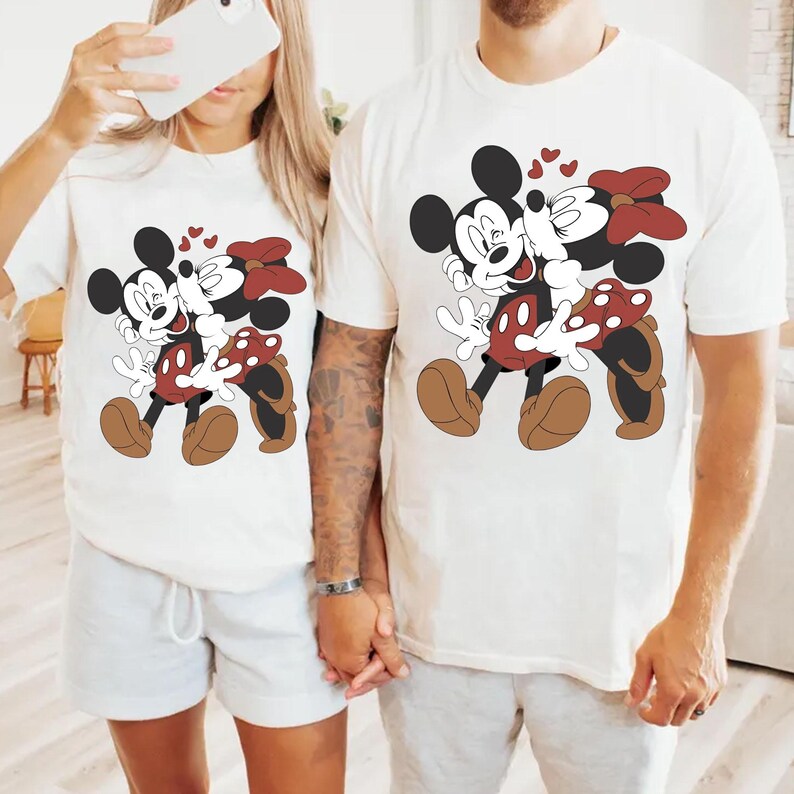 May include: White t-shirts featuring a cartoon image of Mickey and Minnie Mouse embracing, with hearts floating above them. The characters are in their classic outfits, Mickey in red shorts and Minnie in a red polka-dot skirt and bow.