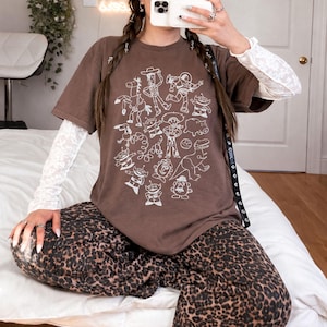 May include: A brown t-shirt featuring a white graphic of cartoon characters. The shirt is paired with a white long-sleeve top and leopard print pants. The overall style is casual and comfortable.