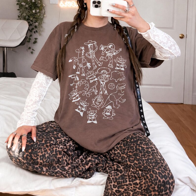 May include: A brown t-shirt featuring a white graphic of cartoon characters. The shirt is paired with a white long-sleeve top and leopard print pants. The overall style is casual and comfortable.