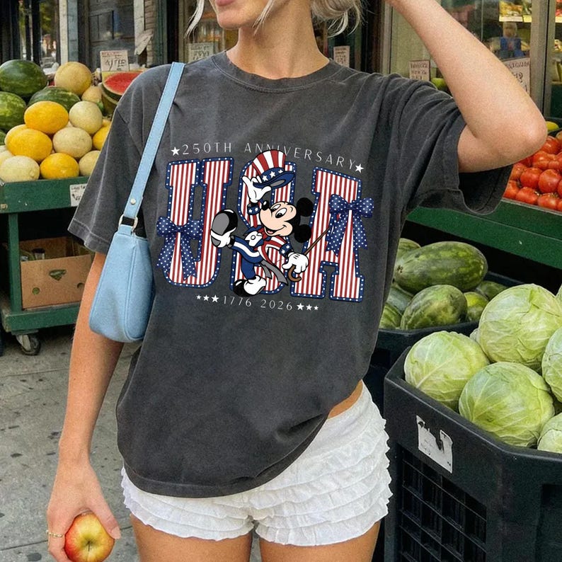 May include: Charcoal grey t-shirt with a patriotic design. The design includes the letters "USA" in red, white, and blue stripes, and a cartoon character in patriotic clothing. The shirt celebrates the 250th anniversary, with the years 1776 and 2026.