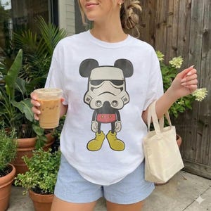May include: White t-shirt with a cartoon design combining Mickey Mouse and a Stormtrooper helmet. The graphic uses black, white, red, and yellow. The shirt is worn with blue shorts. A small canvas tote bag and a drink are also visible.