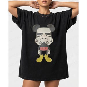 May include: Black t-shirt with a cartoon character mashup design. The graphic combines Mickey Mouse's silhouette with a Stormtrooper helmet. The character has yellow shoes, red shorts, and black ears. The t-shirt has a relaxed fit.
