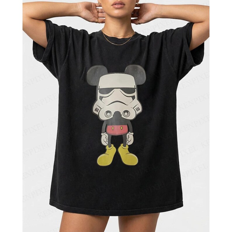 May include: Black t-shirt with a cartoon character mashup design. The graphic combines Mickey Mouse's silhouette with a Stormtrooper helmet. The character has yellow shoes, red shorts, and black ears. The t-shirt has a relaxed fit.