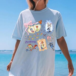 May include: Light blue t-shirt featuring Zootopia characters and the word "Zootopia". The design includes framed images of characters, a pink bow, and decorative elements. The shirt is a light blue color.