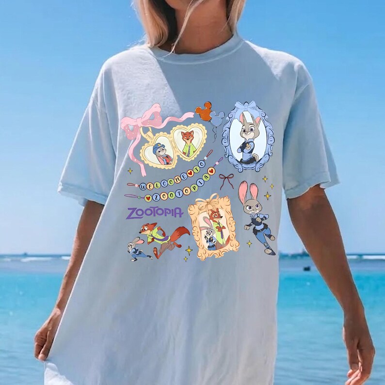 May include: Light blue t-shirt featuring Zootopia characters and the word "Zootopia". The design includes framed images of characters, a pink bow, and decorative elements. The shirt is a light blue color.