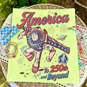 May include: A lime green t-shirt featuring a graphic of a space ranger character in a red, white, and blue suit. The text reads America To 250th and Beyond, accompanied by star, planet, and firework illustrations in red, blue, and dark brown.