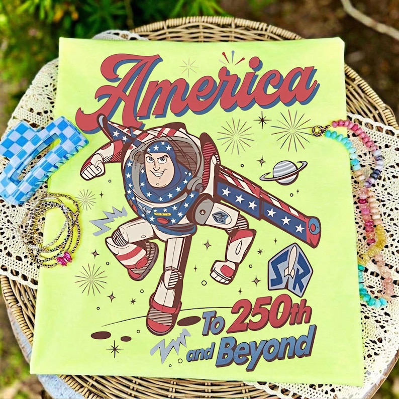 May include: A lime green t-shirt featuring a graphic of a space ranger character in a red, white, and blue suit. The text reads America To 250th and Beyond, accompanied by star, planet, and firework illustrations in red, blue, and dark brown.