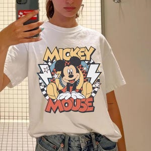 May include: White t-shirt featuring a colorful graphic of Mickey Mouse. The design includes the words "Mickey Mouse" in a retro font, with Mickey sitting in the center. The shirt has short sleeves and a classic crew neck.