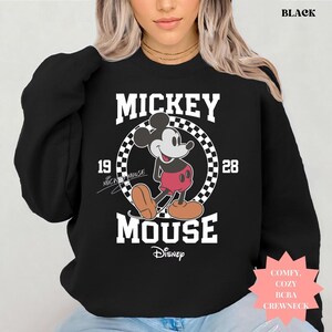 Peut inclure: Sweat-shirt noir &agrave; col rond avec un motif Mickey Mouse. Le design comprend le mot "Mickey" au-dessus d'un motif en damier circulaire avec "1928" de chaque c&ocirc;t&eacute;. Le logo Disney est en bas. Le mot "BLACK" est en haut.