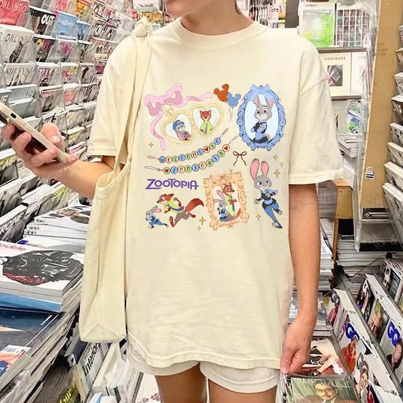 May include: Cream-colored t-shirt featuring various illustrations and the word "Zootopia". The design includes cartoon characters in frames, a string of beads, and a bow. The shirt is a casual wear item.