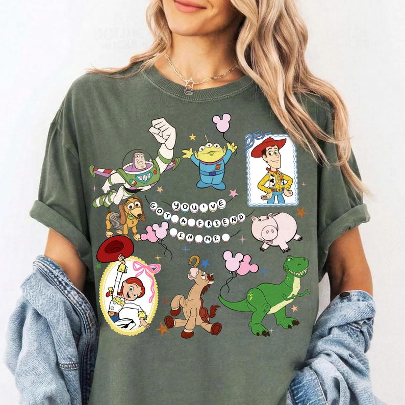 May include: A dark olive green t-shirt featuring a graphic print of various Toy Story characters including Buzz Lightyear, Woody, Jessie, Rex, Slinky Dog, Hamm, and an alien. The text You've Got A Friend In Me is displayed in a circular bead pattern.