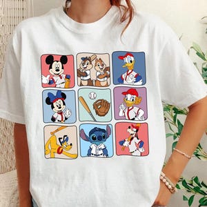 May include: White t-shirt featuring a grid of cartoon characters in baseball uniforms. Mickey Mouse, Chip and Dale, Donald Duck, Minnie Mouse, a baseball, glove, Daisy Duck, Pluto, and Stitch are depicted in colorful squares.