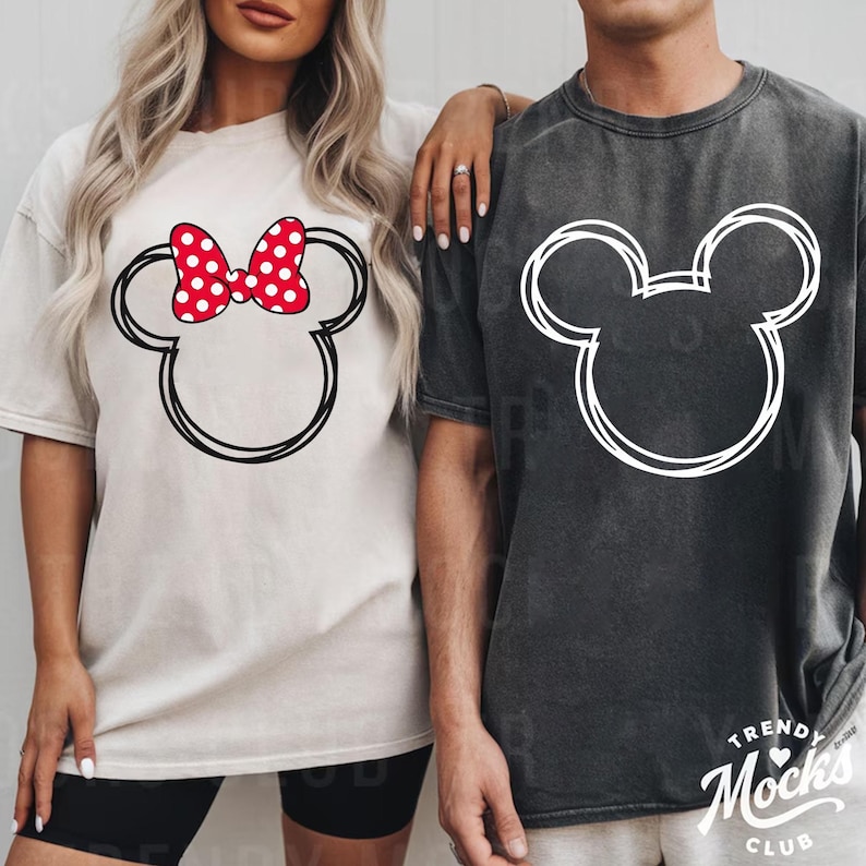 May include: Two oversized t-shirts, one cream and one charcoal, each featuring a Mickey Mouse head outline. The cream shirt has a red polka-dot bow. The charcoal shirt has a white outline. The shirts are part of a matching set.