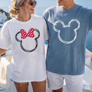 May include: White t-shirt with a black outline of a mouse head and a red polka dot bow. A blue t-shirt with a white mouse head outline is also shown. The shirts are part of a matching set.