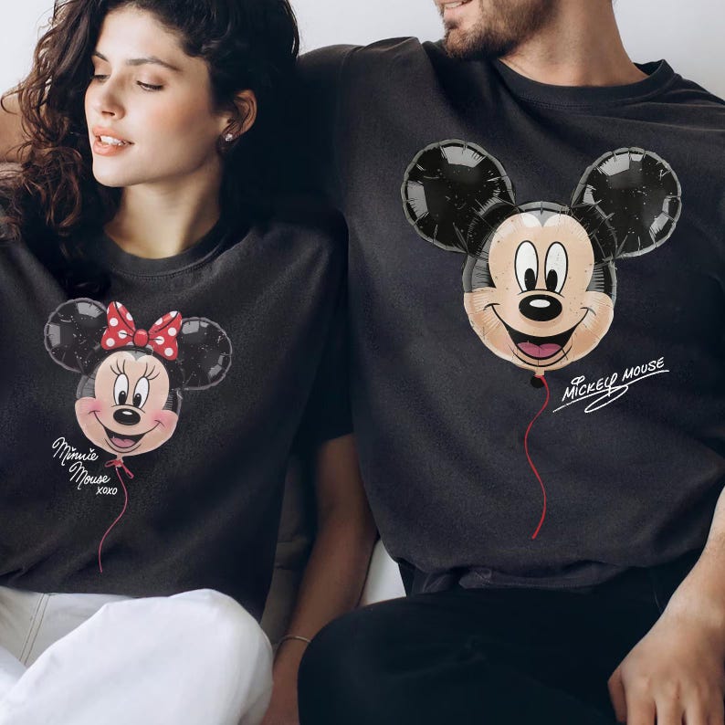 May include: Two dark gray t-shirts featuring balloon-style graphics. One shirt displays a Minnie Mouse balloon with a red polka-dot bow and the text Minnie Mouse xoxo. The other shirt shows a Mickey Mouse balloon with the text Mickey Mouse.