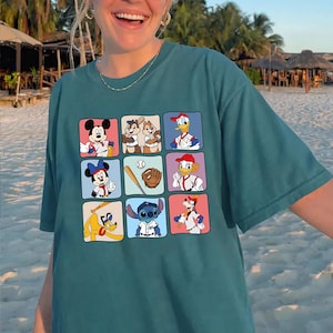May include: Teal-colored t-shirt featuring a grid of cartoon characters in baseball-themed poses. Mickey Mouse, Minnie Mouse, Donald Duck, and Goofy are among the characters. The shirt is a relaxed fit.