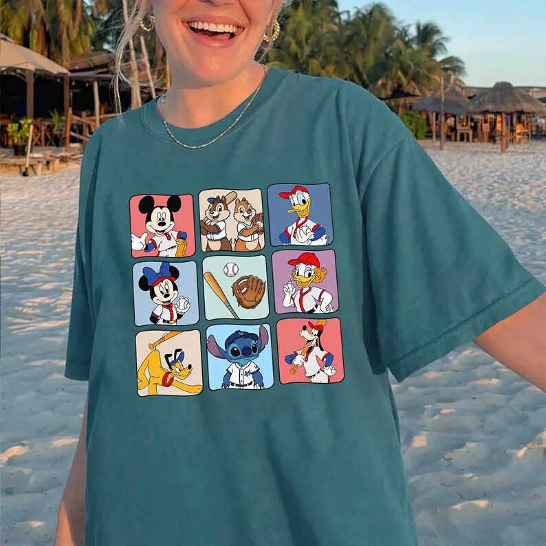 May include: Teal-colored t-shirt featuring a grid of cartoon characters in baseball-themed poses. Mickey Mouse, Minnie Mouse, Donald Duck, and Goofy are among the characters. The shirt is a relaxed fit.