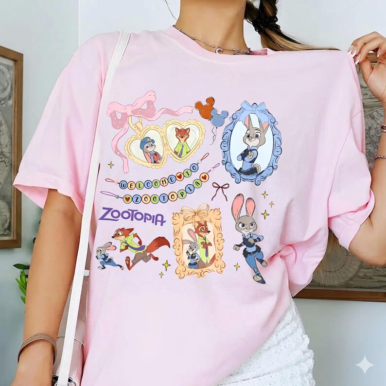 May include: A light pink t-shirt featuring cartoon illustrations from the movie Zootopia. The design includes framed character portraits, the movie's logo, and the phrase "Welcome to Zootopia" in a colorful banner. The shirt has short sleeves.