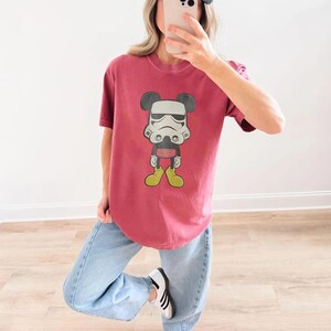 May include: A maroon t-shirt with a graphic of Mickey Mouse in a Stormtrooper helmet. The cartoon character has black ears, a white helmet, red shorts, and yellow shoes. The shirt is worn with light blue jeans and white trainers.