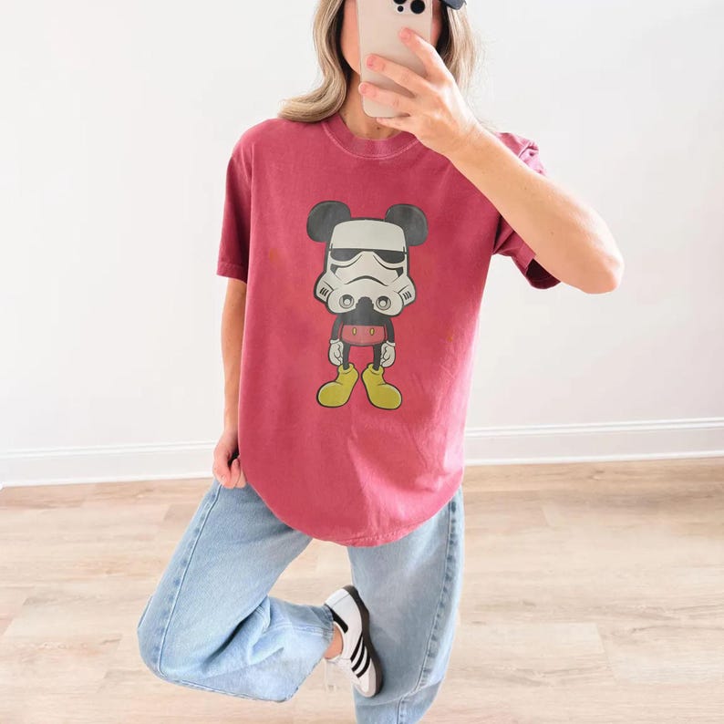 May include: A maroon t-shirt with a graphic of Mickey Mouse in a Stormtrooper helmet. The cartoon character has black ears, a white helmet, red shorts, and yellow shoes. The shirt is worn with light blue jeans and white trainers.