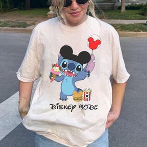 May include: Beige t-shirt featuring a cartoon Stitch character holding an ice cream cone and a Mickey Mouse balloon. The shirt also displays a burger, popcorn, and the words "DISNEY MODE" in black.