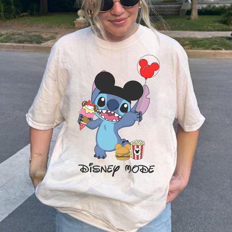 May include: Beige t-shirt featuring a cartoon Stitch character holding an ice cream cone and a Mickey Mouse balloon. The shirt also displays a burger, popcorn, and the words "DISNEY MODE" in black.