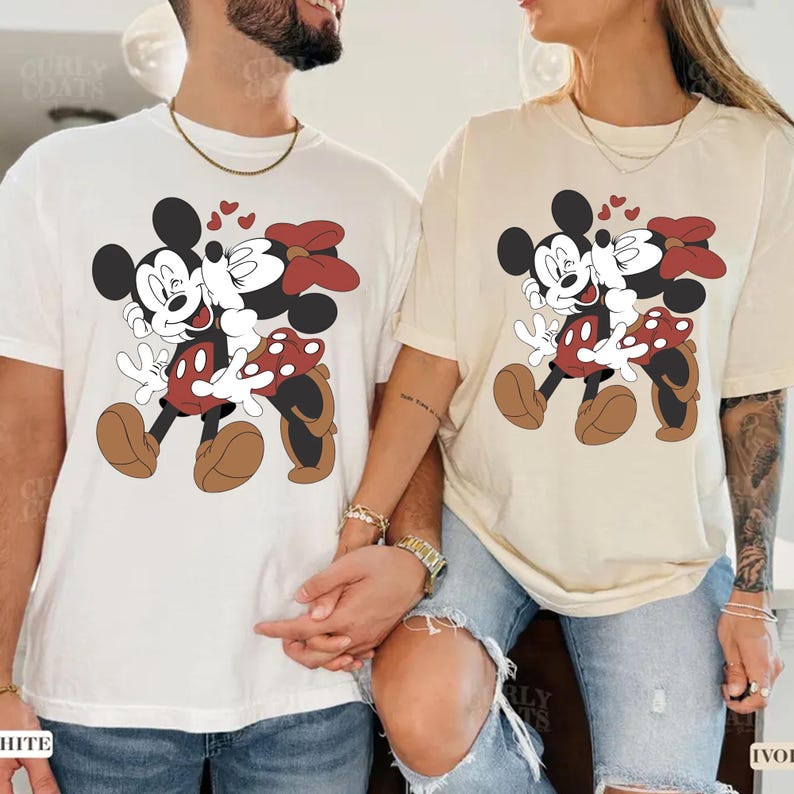 May include: Two t-shirts, one white and one ivory, each featuring a cartoon image of Mickey and Minnie Mouse embracing. Mickey is wearing red shorts with white polka dots, and Minnie has a red bow. Small red hearts float above them.