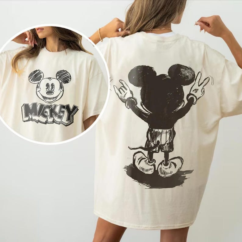 May include: Cream-colored t-shirt featuring a black and white Mickey Mouse graphic. The front of the shirt displays Mickey's face with the word "Mickey" below. The back of the shirt shows a full-body illustration of Mickey Mouse with his arms raised.