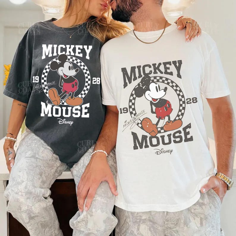 Peut inclure: Deux t-shirts avec un motif Mickey Mouse. L'un est gris fonc&eacute;, l'autre est blanc. Le motif comprend les mots "MICKEY MOUSE" et les ann&eacute;es "19" et "28", avec un cercle &agrave; carreaux autour de Mickey Mouse. Le logo Disney est en bas.