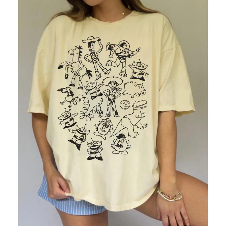 May include: Cream-colored t-shirt featuring a black line drawing of various characters. The graphic includes a cowboy, a space ranger, aliens, and other cartoon figures. The shirt has short sleeves and a relaxed fit.