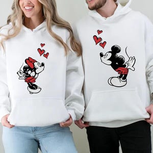May include: White hoodies with Mickey and Minnie Mouse designs. Mickey is in black, red, and white, blowing kisses with red hearts. Minnie is in a red dress and bow, also blowing kisses with red hearts. The hoodies have a front pocket.