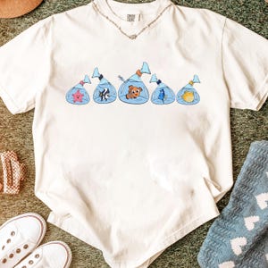May include: Cream-colored t-shirt featuring a cartoon graphic of various fish in plastic bags. The shirt has a crew neck and short sleeves. A silver necklace is visible at the neckline. The shirt is on a green surface.