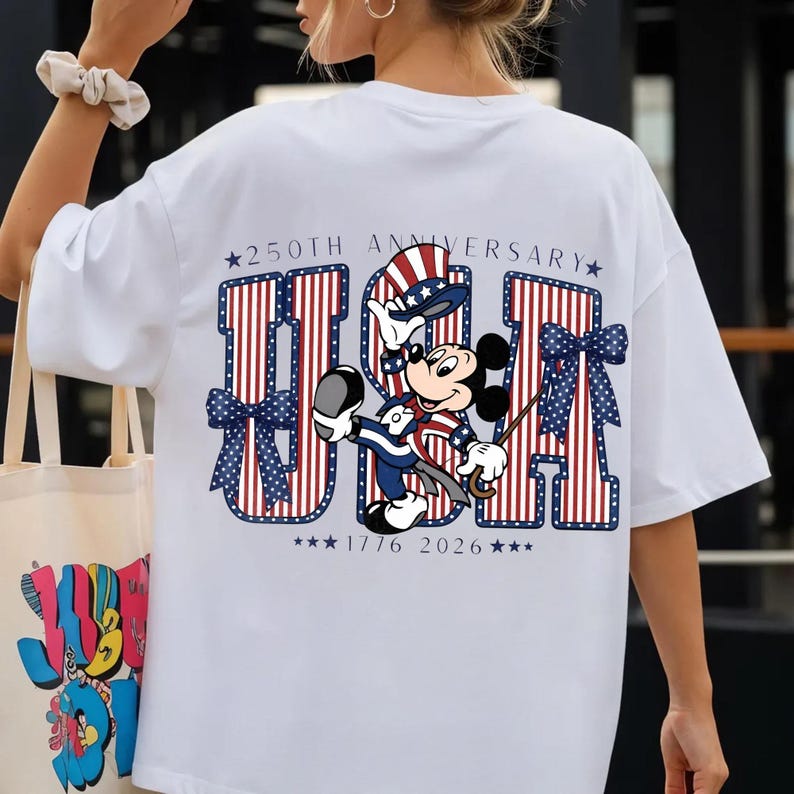 May include: White t-shirt with a patriotic design featuring Mickey Mouse in red, white, and blue. The shirt displays the letters "USA" with stars and stripes, and the text "250th Anniversary 1776 2026".