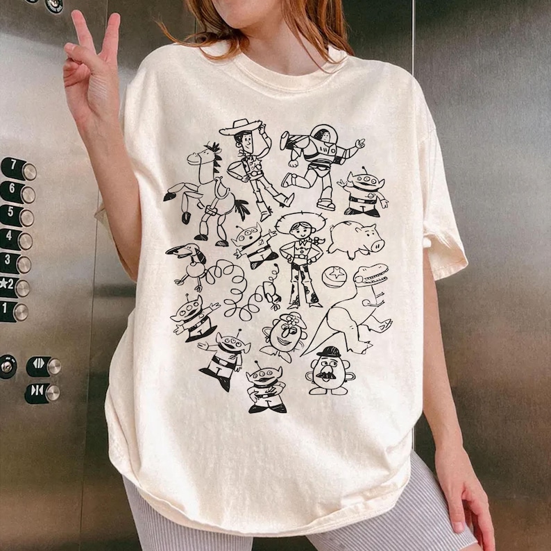May include: Cream-colored t-shirt featuring a black and white illustration of characters from a popular animated movie. The design includes a cowboy, space ranger, and other recognizable figures. The shirt has a relaxed fit.