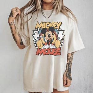 May include: Cream-colored t-shirt featuring a graphic of Mickey Mouse. The design includes the words "Mickey Mouse" in a retro font, with Mickey in the center. The graphic is surrounded by stars, lightning bolts, and a checkered pattern.