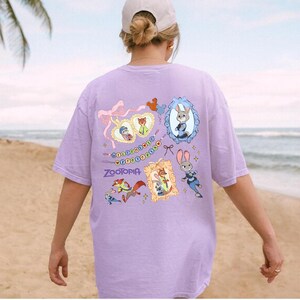 May include: A lilac-colored t-shirt featuring cartoon characters and the word "Zootopia." The design includes framed portraits of characters, a heart-shaped design, and a string of beads. The shirt is worn by a person on a beach.