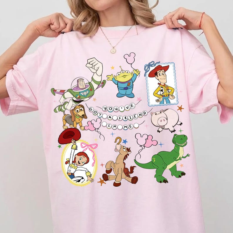 May include: A light pink t-shirt featuring a graphic print of Toy Story characters including Buzz Lightyear, Woody, Jessie, Rex, Slinky Dog, Hamm, Bullseye, and an Alien. The text "YOU'VE GOT A FRIEND IN ME" is displayed in the center with small balloons.