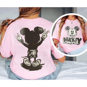 May include: Pink t-shirt featuring a black and white graphic of Mickey Mouse. The back of the shirt shows Mickey Mouse with his hands in a rock and roll gesture. The front of the shirt has a Mickey Mouse face and the word "Mickey".