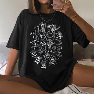 May include: Black t-shirt featuring a white graphic of various characters from a popular animated movie. The design includes a cowboy, spaceman, and other recognizable figures. The shirt is a casual, oversized style.