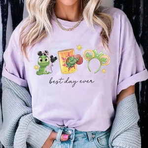 Op de afbeelding: Lichtpaars T-shirt met een cartoonafbeelding van een groene kameleon, een lantaarn en een hoofdband met oren. De tekst "best day ever" staat in zwart script eronder.