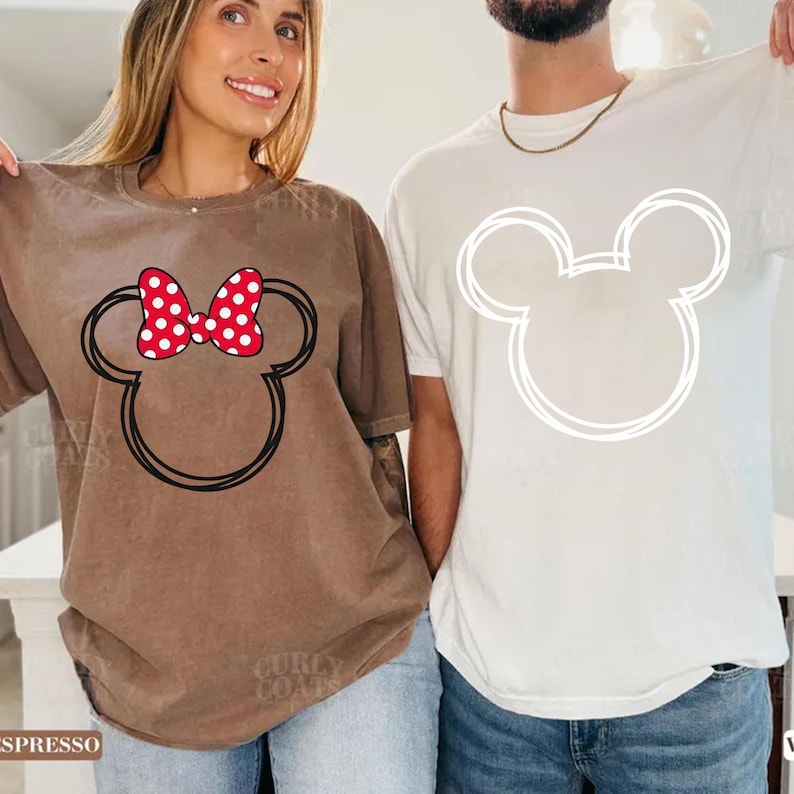 May include: Two matching t-shirts. One is espresso-colored with a black outline of Mickey Mouse's head and a red polka-dot bow. The other is white with a white outline of Mickey Mouse's head. Both shirts are worn by people.